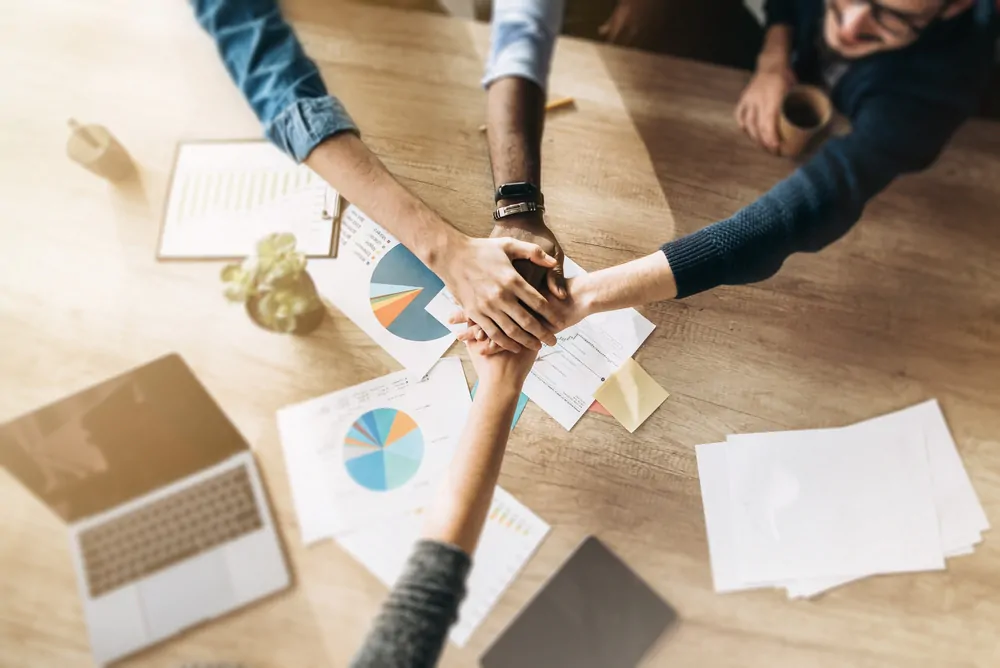 Pessoas colocando a mão uma por cima da outra com papéis e computador em baixo.
